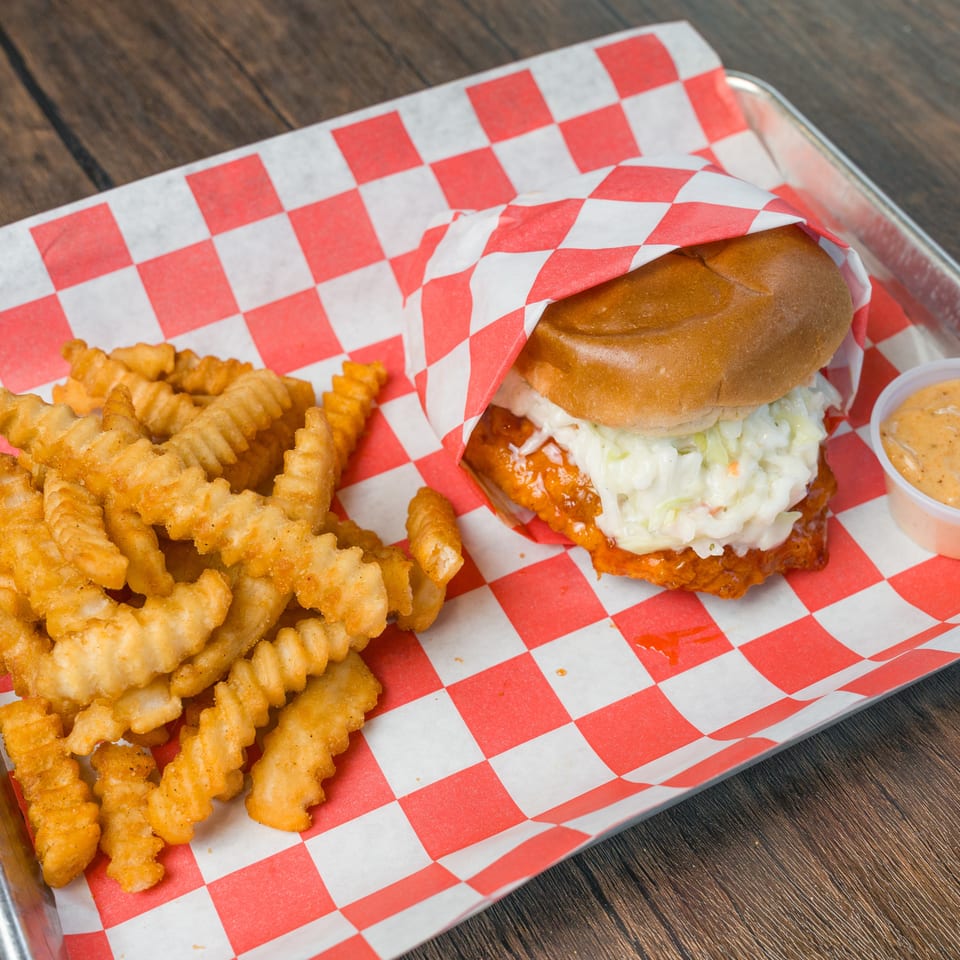Fried chicken sandwich with coleslaw, crinkle fries, and dipping sauce on a checkered paper-lined tray.
