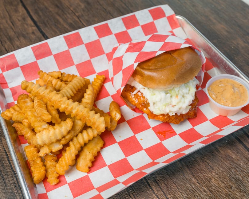Fried chicken sandwich with coleslaw, crinkle fries, and dipping sauce on a checkered paper-lined tray.