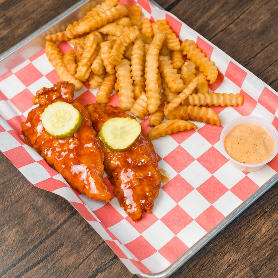 Two saucy chicken tenders with pickles, crinkle fries, and dipping sauce on a checkered tray.