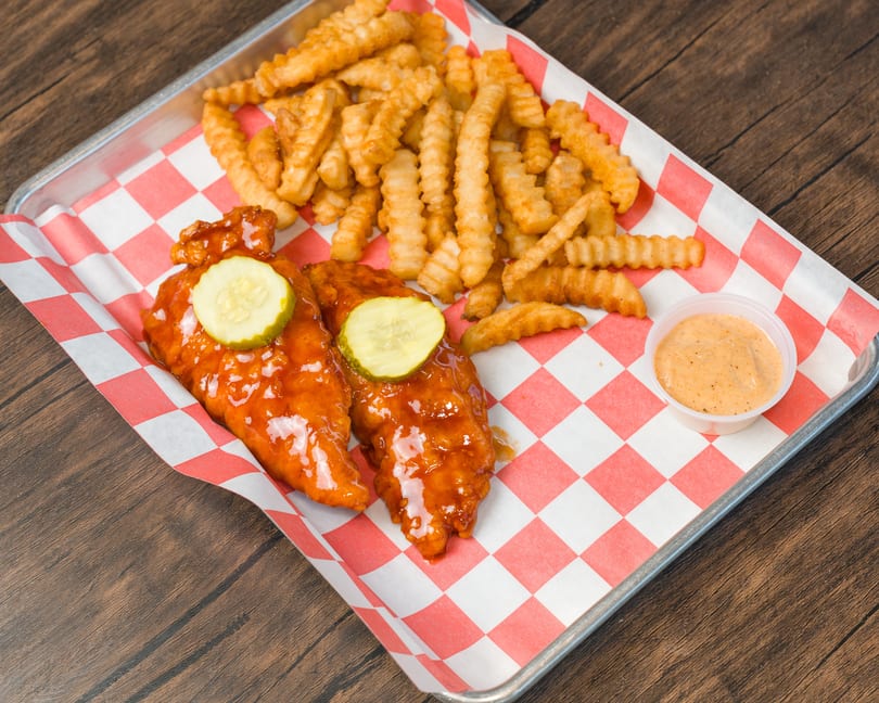 Two saucy chicken tenders with pickles, crinkle fries, and dipping sauce on a checkered tray.