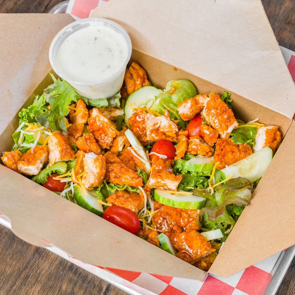 Buffalo chicken salad with lettuce, tomatoes, cucumbers, cheese, and ranch dressing in a takeout box.
