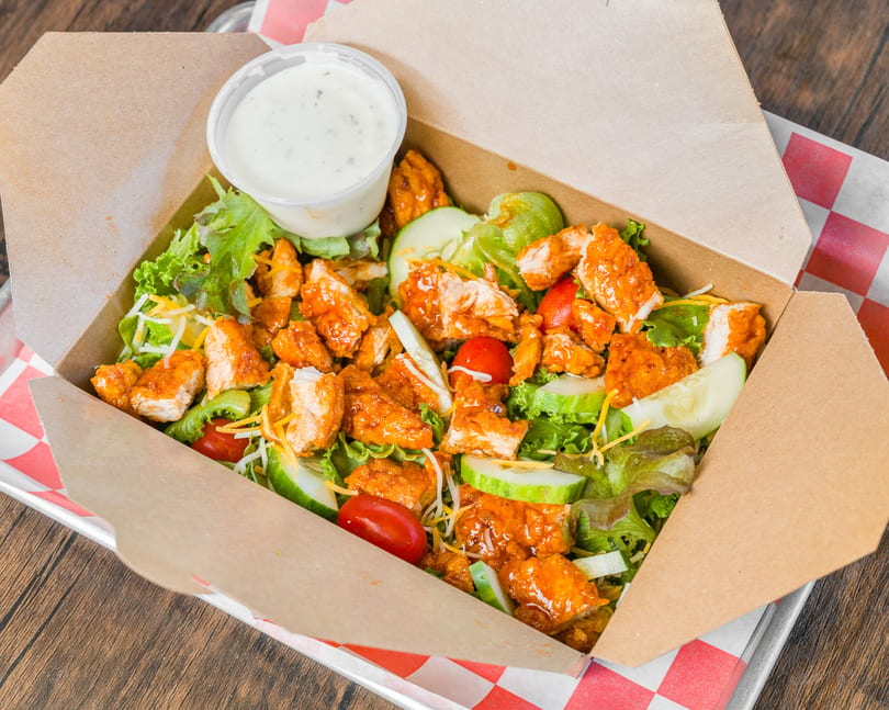 Buffalo chicken salad with lettuce, tomatoes, cucumbers, cheese, and ranch dressing in a takeout box.