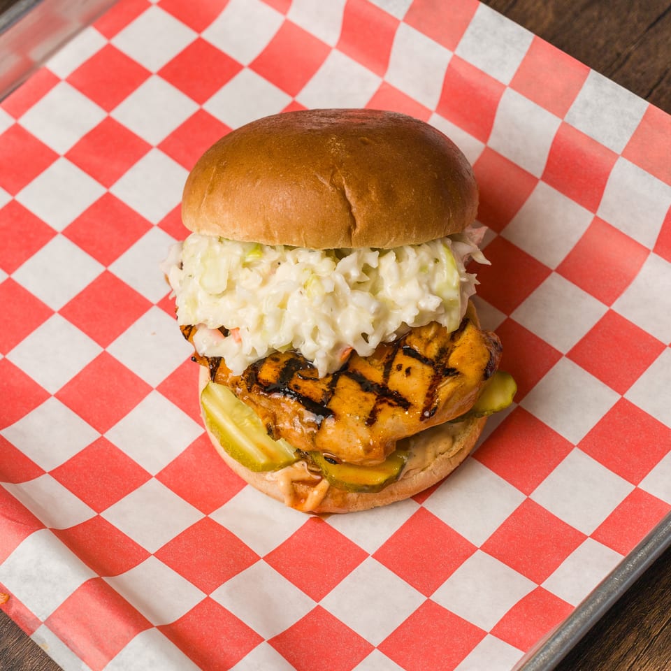Grilled chicken sandwich with char marks, coleslaw, and pickles on red checkered paper.