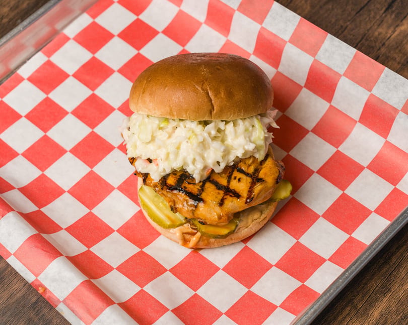 Grilled chicken sandwich with char marks, coleslaw, and pickles on red checkered paper.