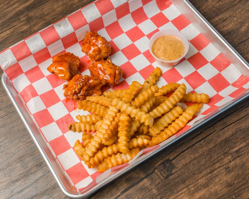 Boneless chicken wings, crinkle-cut fries, and dipping sauce on a metal tray with checkered paper.